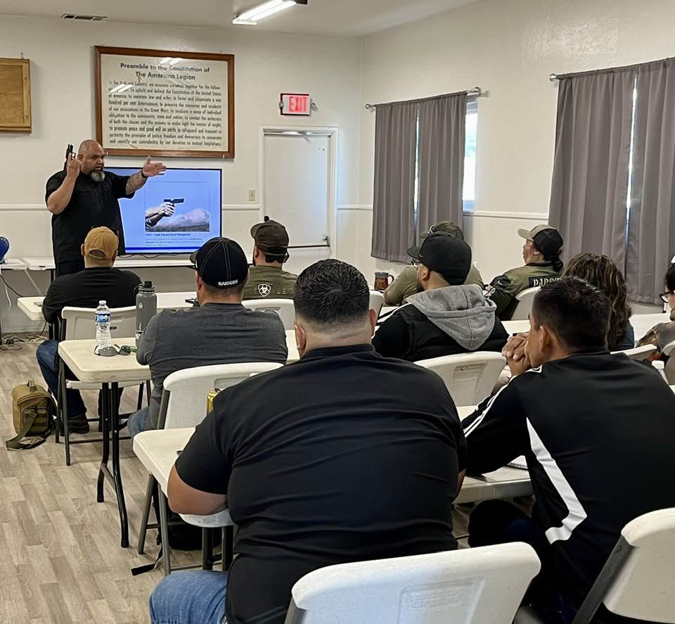 This image captures an instructor actively engaging with students during an introduction to firearms class. The instructor is demonstrating a technique or safety principle related to handgun handling, while the students attentively observe and take notes. The classroom setting includes a screen displaying an instructional slide, tables, and chairs arranged to facilitate learning, and the presence of various students indicating an interactive, group learning environment. This image highlights the practical and educational aspects of the class, emphasizing hands-on instruction and focused attention on firearm safety and handling skills.